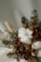A detailed arrangement of dried flowers and plants, including green pine branches, brown leaves, fluffy white cotton, and small dried yellow buds. The natural textures and colors blend gently, suggesting a rustic and organic aesthetic.