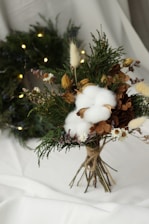 A bouquet made of a variety of natural elements including cotton bolls, dried flowers, greenery, and brown foliage, tied together with twine. It is arranged on a white surface with a wreath adorned with small lights visible in the background.