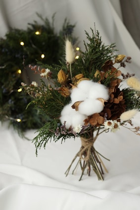 A bouquet made of a variety of natural elements including cotton bolls, dried flowers, greenery, and brown foliage, tied together with twine. It is arranged on a white surface with a wreath adorned with small lights visible in the background.
