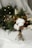 A bouquet made of a variety of natural elements including cotton bolls, dried flowers, greenery, and brown foliage, tied together with twine. It is arranged on a white surface with a wreath adorned with small lights visible in the background.