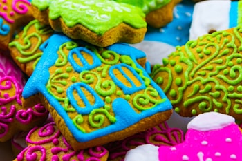 Brightly colored cookies with intricate frosting designs featuring swirls and patterns in blue, green, pink, and other vibrant hues. One notable cookie is shaped like a house with detailed window designs.