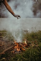A person holds a glass jar over a small, burning fire surrounded by grass and twigs. Smoke rises through the air, and the scene is set outdoors, possibly indicating an extinguishing act or a controlled fire.
