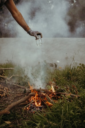A person holds a glass jar over a small, burning fire surrounded by grass and twigs. Smoke rises through the air, and the scene is set outdoors, possibly indicating an extinguishing act or a controlled fire.
