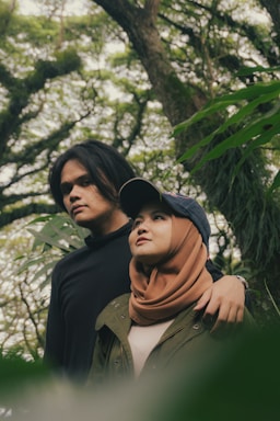 A couple stands together in a lush, green forest setting. The pair appears relaxed and contemplative, with one wearing a brown hijab and the other dressed in a black top. The surrounding trees and foliage create a serene and natural backdrop.