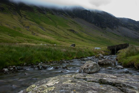 A serene landscape featuring a small, flowing stream bordered by rocks in the foreground. The background reveals rolling green hills and a dark, rocky hillside partially obscured by low, misty clouds. A rustic stone bridge or drainage structure is visible to the right.