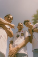Three young individuals wearing school uniforms are standing outdoors, looking downwards. The sky is clear and blue with the presence of a palm tree on the right side of the image. The photograph has a warm and nostalgic feel due to the soft focus and lighting.