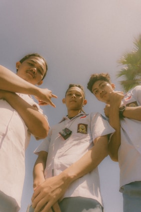 Three young individuals wearing school uniforms are standing outdoors, looking downwards. The sky is clear and blue with the presence of a palm tree on the right side of the image. The photograph has a warm and nostalgic feel due to the soft focus and lighting.