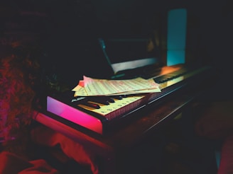 A keyboard sits on a wooden surface, partially covered by sheet music. The scene is dimly lit, creating a moody and intimate atmosphere. Soft ambient lighting in hues of pink and blue highlights the keyboard and music sheets.
