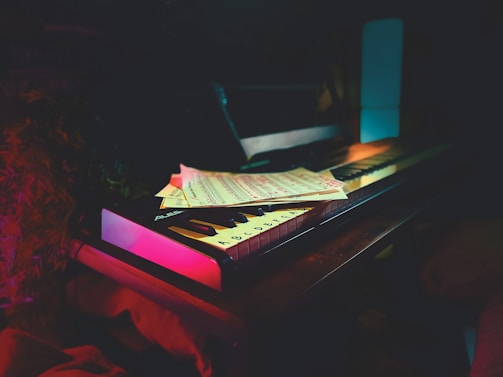 A keyboard sits on a wooden surface, partially covered by sheet music. The scene is dimly lit, creating a moody and intimate atmosphere. Soft ambient lighting in hues of pink and blue highlights the keyboard and music sheets.