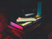 A keyboard sits on a wooden surface, partially covered by sheet music. The scene is dimly lit, creating a moody and intimate atmosphere. Soft ambient lighting in hues of pink and blue highlights the keyboard and music sheets.