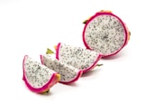 Several pieces of dragon fruit are arranged on a plain white background. The fruit shows a vibrant pink outer skin with a speckled white flesh that has tiny black seeds. One piece is fully intact, while the others are cut into wedges.