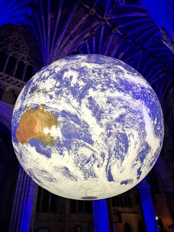 A large, detailed globe depicting Earth is suspended in an indoor setting with an ornate ceiling. The globe prominently features the continents and cloud patterns, illuminated with a mix of blue and white lighting. The surroundings are dimly lit, with architectural elements visible in the background.