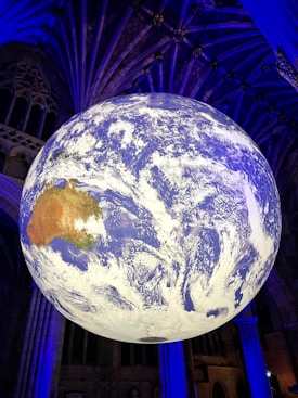 A large, detailed globe depicting Earth is suspended in an indoor setting with an ornate ceiling. The globe prominently features the continents and cloud patterns, illuminated with a mix of blue and white lighting. The surroundings are dimly lit, with architectural elements visible in the background.