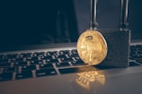 A golden Bitcoin coin is secured with a strong black padlock placed on top of a laptop keyboard with Russian layout. The image symbolizes security and protection in the context of digital currency.