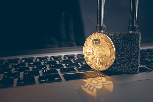 A golden Bitcoin coin is secured with a strong black padlock placed on top of a laptop keyboard with Russian layout. The image symbolizes security and protection in the context of digital currency.