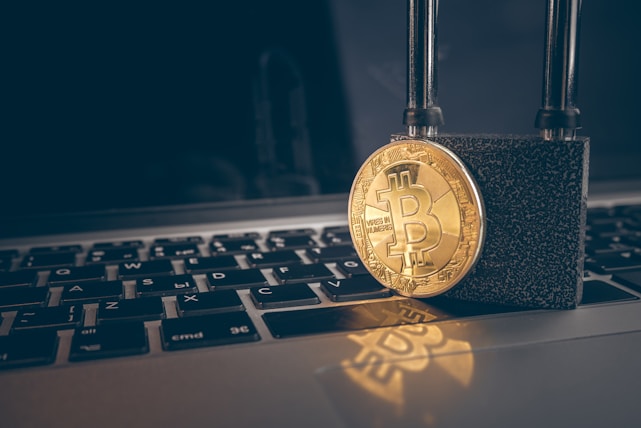 A golden Bitcoin coin is secured with a strong black padlock placed on top of a laptop keyboard with Russian layout. The image symbolizes security and protection in the context of digital currency.