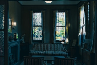 A dimly lit, vintage-style living room with a striped sofa in front of three large windows featuring stained glass. Long, elegant curtains frame the windows, while soft light illuminates the room, highlighting a fireplace and decorative items like a statue and lamp. The atmosphere exudes a sense of nostalgia and tranquility.