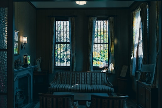 A dimly lit, vintage-style living room with a striped sofa in front of three large windows featuring stained glass. Long, elegant curtains frame the windows, while soft light illuminates the room, highlighting a fireplace and decorative items like a statue and lamp. The atmosphere exudes a sense of nostalgia and tranquility.