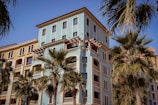 A Mediterranean-style apartment building with a pale blue facade and decorative arches, surrounded by palm trees under a clear blue sky.