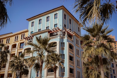 A Mediterranean-style apartment building with a pale blue facade and decorative arches, surrounded by palm trees under a clear blue sky.