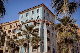 A Mediterranean-style apartment building with a pale blue facade and decorative arches, surrounded by palm trees under a clear blue sky.