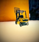 A small, detailed model of a yellow forklift is placed on a smooth, reflective surface. The background features a gradient of warm tones with a blend of dark and light areas, creating a vivid contrast with the model's color.