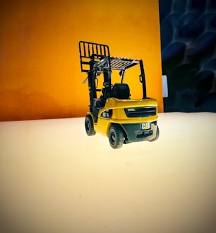 A small, detailed model of a yellow forklift is placed on a smooth, reflective surface. The background features a gradient of warm tones with a blend of dark and light areas, creating a vivid contrast with the model's color.