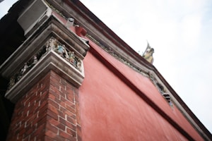 A detailed view of a building corner featuring intricate carvings and a combination of brick and plaster surfaces. The architecture includes ornate sculptures near the roofline and a CCTV camera mounted at the corner.