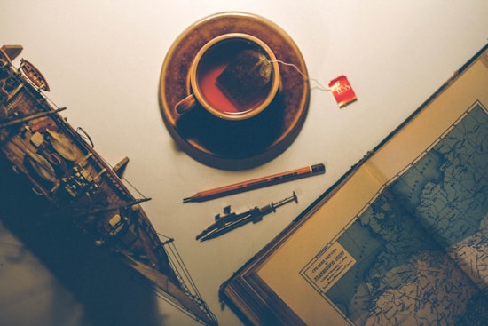 A vintage tabletop scene featuring a cup of tea with a teabag labeled 'TKS', a detailed model ship, a mechanical pencil, and an open book showcasing a map. The lighting is warm and creates a nostalgic ambiance.