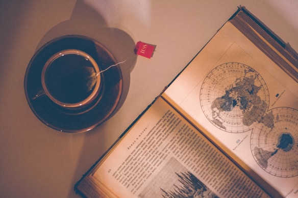A vintage-style photograph featuring a cup of tea placed on a saucer with a dangling tea bag. Next to it lies an open book displaying maps, possibly indicating historical or geographical content. The warm color palette and lighting create a nostalgic atmosphere.