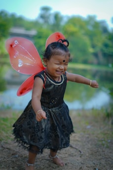 A young child wearing a black dress with floral patterns is joyfully playing outdoors. The child has red butterfly wings attached, standing on a grassy area near a pond surrounded by lush greenery. The child is smiling with delight, and the expression conveys a sense of carefree happiness.