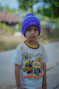 A child is wearing a colorful t-shirt featuring cartoon characters and a purple knit cap. The background shows blurred greenery and a rustic structure, suggesting an outdoor setting.