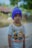 A child is wearing a colorful t-shirt featuring cartoon characters and a purple knit cap. The background shows blurred greenery and a rustic structure, suggesting an outdoor setting.