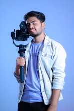 A young man is holding a camera mounted on a stabilizer. He is wearing a light denim jacket over a lavender t-shirt, standing against a solid blue background. The man's eyes are closed, and he seems to be in a relaxed, content state.