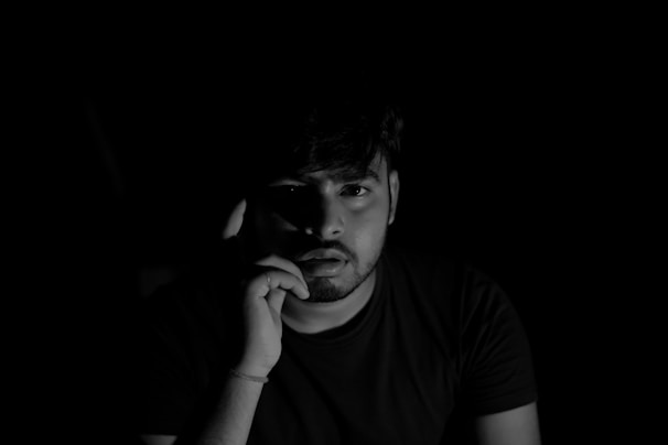 A person sits in a dark environment, partially illuminated from the side, creating a dramatic shadow effect. The individual appears to be in thought with a hand resting on their face. The black and white composition enhances the contrast between light and shadow.