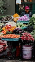 A vibrant market stall displaying an array of fresh vegetables including carrots, red radishes, various peppers, eggplants, and cabbages. A person wearing a red cap and a jacket stands behind the colorful display. Piles of purple onions, root vegetables, and neatly arranged produce are visible. The stall is set against a dark green background, with some leafy plants visible on the side.