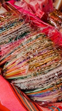 A collection of colorful, striped candy sticks or sweets is wrapped in transparent plastic. The candies are arranged in stacks, and are surrounded by bright, vibrant textiles.
