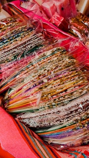 A collection of colorful, striped candy sticks or sweets is wrapped in transparent plastic. The candies are arranged in stacks, and are surrounded by bright, vibrant textiles.