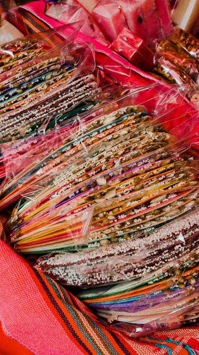 A collection of colorful, striped candy sticks or sweets is wrapped in transparent plastic. The candies are arranged in stacks, and are surrounded by bright, vibrant textiles.