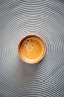 A small cup filled with espresso sits on a surface with concentric circular grooves, creating a visually appealing pattern. The espresso has a creamy, frothy texture with a few bubbles visible on the surface.