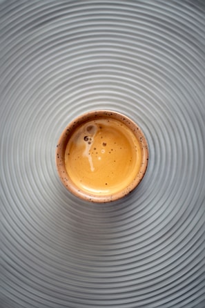 A small cup filled with espresso sits on a surface with concentric circular grooves, creating a visually appealing pattern. The espresso has a creamy, frothy texture with a few bubbles visible on the surface.