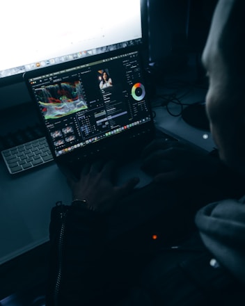 A person is sitting at a desk with a laptop open, displaying video editing software. The screen shows color correction tools and a preview of a video on the editing timeline. The room is dimly lit, creating a focused and immersive atmosphere.