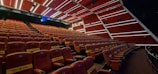 A cinema hall with rows of empty red seats, featuring a modern design with diagonal white lines on the walls and ceiling. The seats have cup holders and plush padding, and some are marked with reservation signs. The lighting is dim, with some ambient lights illuminating the space.