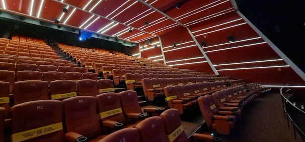 A cinema hall with rows of empty red seats, featuring a modern design with diagonal white lines on the walls and ceiling. The seats have cup holders and plush padding, and some are marked with reservation signs. The lighting is dim, with some ambient lights illuminating the space.