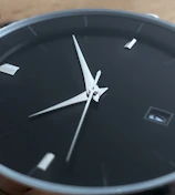 Close-up of a sleek black luxury watch face with silver detailing on a minimalist white background.