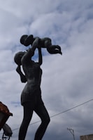 A bronze statue depicting a woman holding a small child above her head against a cloudy sky. The woman is dressed in shorts and has her hair tied back, displaying a sense of joy and care.