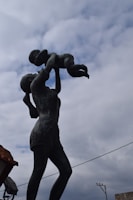 A bronze statue depicting a woman holding a small child above her head against a cloudy sky. The woman is dressed in shorts and has her hair tied back, displaying a sense of joy and care.
