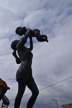 A bronze statue depicting a woman holding a small child above her head against a cloudy sky. The woman is dressed in shorts and has her hair tied back, displaying a sense of joy and care.