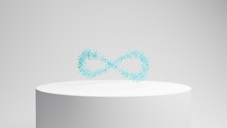 A white cylindrical pedestal displays a floating collection of small, turquoise particles forming the shape of an infinity symbol against a neutral background.