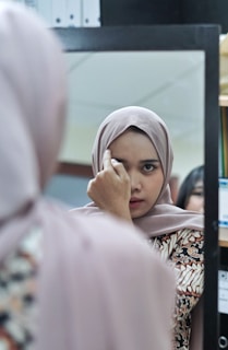 A woman wearing a hijab is looking at herself in a mirror, adjusting her hijab with one hand. The reflection captures her focused expression. She is in an indoor setting, possibly an office or a home, as indicated by the shelves and files in the background.